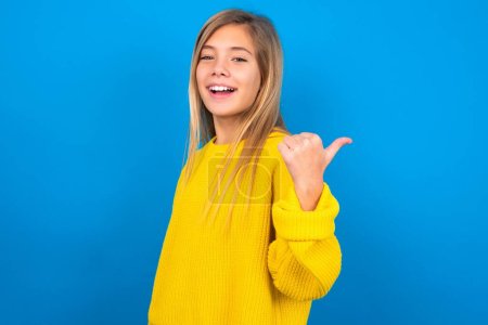 Photo for Impressed caucasian teen girl wearing yellow sweater over blue studio background points back empty space - Royalty Free Image