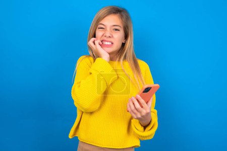 Foto de Miedo caucásico adolescente chica usando amarillo suéter celebración de teléfono y morder las uñas en fondo azul - Imagen libre de derechos