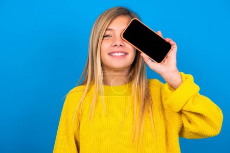 Téléchargez les photos : Belle adolescente caucasienne portant un pull jaune tenant smartphone moderne couvrant un œil tout en souriant et posant sur fond bleu - en image libre de droit