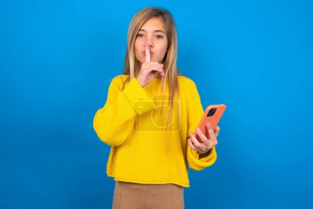 Téléchargez les photos : Blonde caucasien adolescent fille portant pull jaune sur fond bleu studio tenant gadget moderne demander de ne pas dire de secrets - en image libre de droit