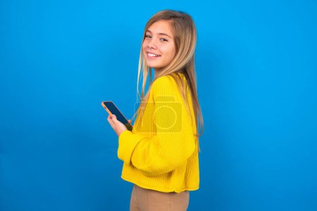 Téléchargez les photos : Vue arrière portrait de caucasienne adolescent fille portant pull jaune sur fond bleu studio en utilisant smartphone souriant - en image libre de droit