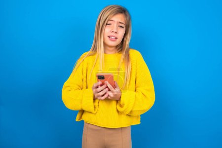 Foto de Foto de chica adolescente caucásica asombrada usando suéter amarillo sobre fondo de estudio azul celebración de teléfono inteligente - Imagen libre de derechos
