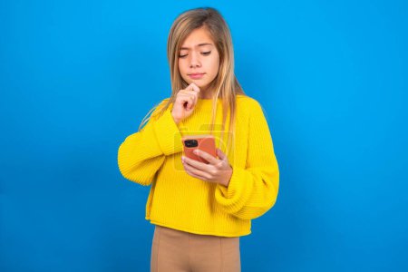 Photo for Thoughtful caucasian teen girl wearing yellow sweater over blue studio background looking at phone - Royalty Free Image