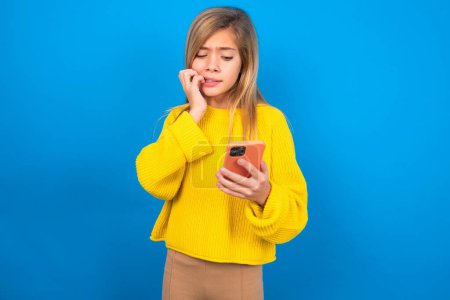 Téléchargez les photos : Portrait d'une adolescente caucasienne assez effrayée portant un pull jaune sur un studio bleu bavardant sur des ongles mordants après avoir lu des nouvelles effrayantes sur son smartphone. - en image libre de droit