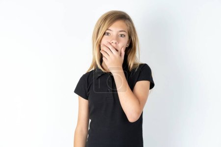 Foto de Emocional hermosa chica adolescente caucásica vistiendo camiseta negra sobre blanco jadeos walll de asombro, cubre la boca abierta con la palma de la mano, se ve sorprendido por la cámara. - Imagen libre de derechos