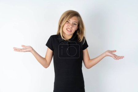 Photo for Clueless beautiful caucasian teen girl wearing black T-shirt over white wall shrugs shoulders with hesitation, faces doubtful situation, spreads palms, Hard decision - Royalty Free Image