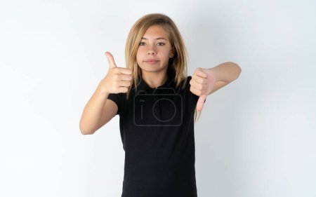 Téléchargez les photos : Belle caucasienne adolescent fille portant noir T-shirt sur blanc mur montrant pouces vers le haut et pouces vers le bas, difficile choisir concept - en image libre de droit