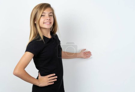 Foto de Hermosa chica adolescente caucásica vistiendo camiseta negra sobre la pared blanca sintiéndose feliz y alegre, sonriendo y dándote la bienvenida, invitándote con un gesto amistoso - Imagen libre de derechos