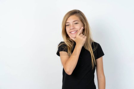 Téléchargez les photos : Belle adolescente caucasienne portant un T-shirt noir sur un mur blanc regardant avec confiance la caméra souriant avec les bras croisés et la main levée sur le menton. Penser positif - en image libre de droit