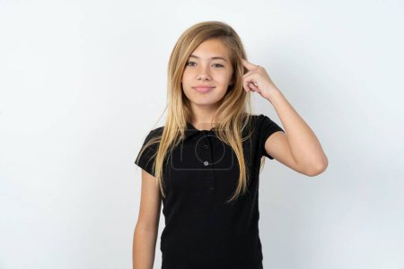Photo for Beautiful caucasian teen girl wearing black T-shirt over white wall tries to memorize something, keeps fore finger on temple, reminds information for exam - Royalty Free Image