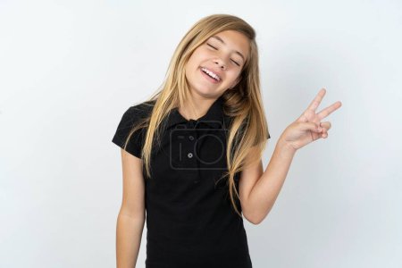 Téléchargez les photos : Belle adolescente caucasienne portant un T-shirt noir sur un mur blanc souriant avec un clin d'oeil heureux au visage de la caméra faisant signe de victoire. Numéro deux - en image libre de droit