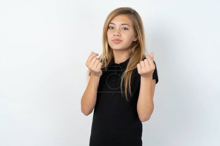 Foto de Hermosa chica adolescente caucásica vistiendo camiseta negra sobre la pared blanca haciendo gesto de dinero con las manos, pidiendo pago de sueldo, negocio millonario - Imagen libre de derechos