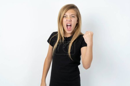 beautiful caucasian teen girl wearing black T-shirt over white wall angry and mad raising fist frustrated and furious while shouting with anger. Rage and aggressive concept