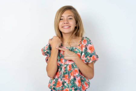 Téléchargez les photos : Belle adolescente caucasienne portant chemisier fleuri sur mur blanc pressé pointant vers la montre-bracelet, impatience, regardant la caméra avec une expression détendue - en image libre de droit
