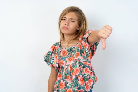 Photo for Beautiful caucasian teen girl wearing flowered blouse over white wall feeling angry, annoyed, disappointed or displeased, showing thumbs down with a serious look - Royalty Free Image