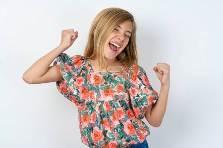 Ecstatic beautiful caucasian teen girl wearing flowered blouse over white wall shout loud yeah fist up raise win lottery