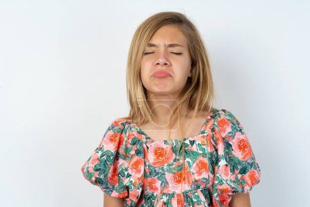 Photo for Beautiful caucasian teen girl wearing flowered blouse over white wall keeps teeth clenched, frowns face in dissatisfaction, irritated because of much duties. - Royalty Free Image