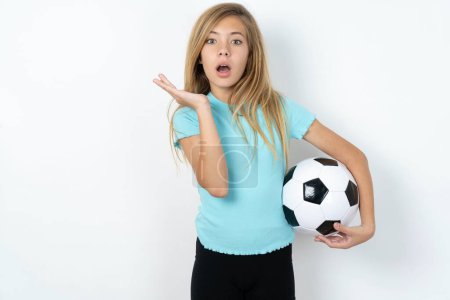Téléchargez les photos : Surpris terrifié caucasien adolescent fille portant des vêtements de sport tenant un ballon de football sur le mur blanc gestes avec incertitude, regarde la caméra, perplexe comme ne sait pas réponse sur la question délicate, Les gens, langage corporel, concept d'émotions - en image libre de droit