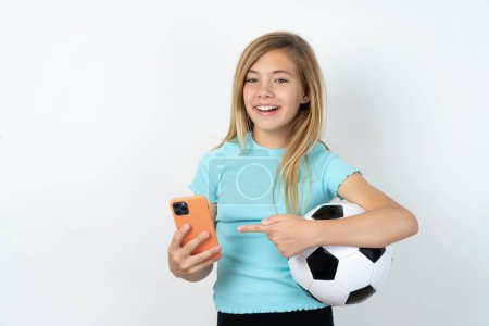 Foto de Chica adolescente caucásica usando ropa deportiva sosteniendo una pelota de fútbol sobre pared blanca sosteniendo en las manos mostrando una nueva celda, - Imagen libre de derechos