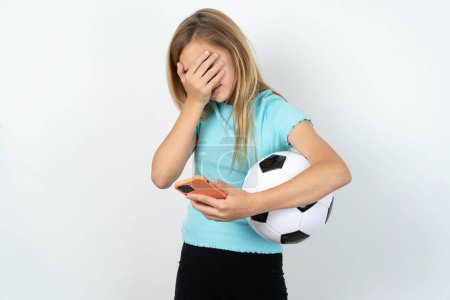 Photo for Caucasian teen girl wearing sportswear holding a football ball over white wall looking at smartphone feeling sad holding hand on face. - Royalty Free Image