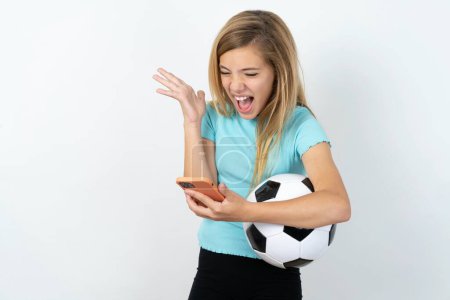 Foto de Enojado hermosa chica adolescente caucásica usando ropa deportiva sosteniendo una pelota de fútbol sobre la pared blanca gritando por teléfono. Problemas en el trabajo. - Imagen libre de derechos