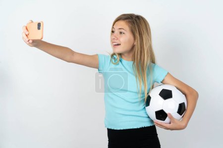 Foto de Hermosa chica adolescente caucásica usando ropa deportiva sosteniendo una pelota de fútbol sobre la pared blanca sonriendo y tomando un selfie listo para publicarlo en sus redes sociales. - Imagen libre de derechos