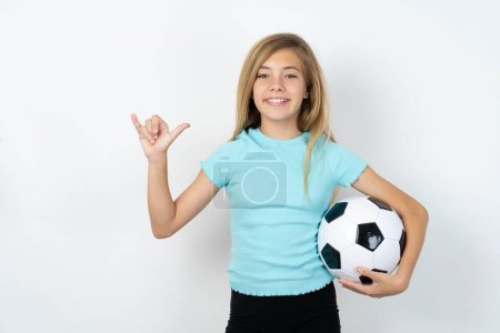Foto de Hermosa chica adolescente caucásica con ropa deportiva sosteniendo una pelota de fútbol sobre la pared blanca que muestra el número seis Liu con el gesto de los dedos en el lenguaje chino signo - Imagen libre de derechos