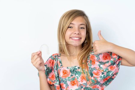 wunderschönes kaukasisches Teenie-Mädchen in geblümter Bluse über der weißen Wand, das eine unsichtbare Zahnspange in der Hand hält und den Daumen nach oben reckt und diese neue Behandlung empfiehlt. Zahnmedizinisches Konzept.