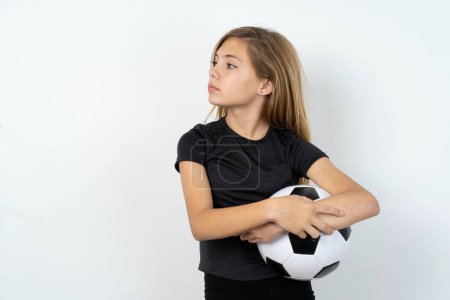 Photo for Charming and thoughtful teen girl wearing sportswear holding a football ball over white wall stands with arms folded concentrated somewhere with pensive expression thinks what to do - Royalty Free Image