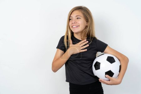 Foto de Jovencita alegre con ropa deportiva sosteniendo una pelota de fútbol sobre la pared blanca expresa emociones positivas recuerda algo divertido mantiene la mano en el pecho y se ríe felizmente. - Imagen libre de derechos