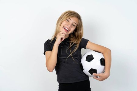 Foto de Hermosa chica adolescente caucásica usando ropa deportiva sosteniendo una pelota de fútbol sobre la pared blanca risas felizmente mantiene la mano en la barbilla expresa emociones positivas sonrisas ampliamente tiene expresión despreocupada - Imagen libre de derechos