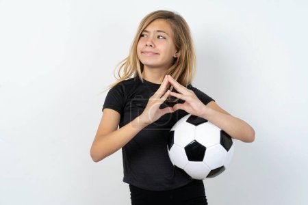 Photo for Teen girl wearing sportswear holding a football ball over white wall steepled fingers and looks mysterious aside has great evil plan in mind - Royalty Free Image