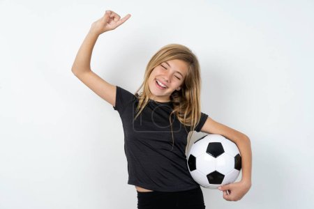 Téléchargez les photos : Photo de l'adolescente optimiste portant des vêtements de sport tenant un ballon de football sur un mur blanc a du plaisir et danse vêtements insouciants être d'humeur parfaite fait des mouvements. Passe du temps libre sur disco partie - en image libre de droit