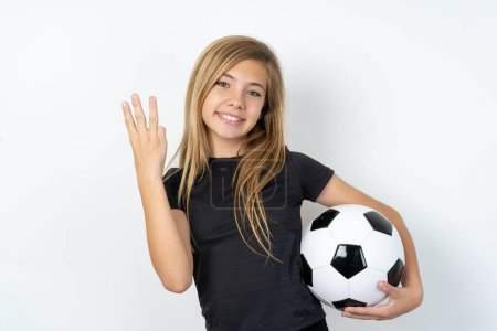 Photo for Teen girl wearing sportswear holding a football ball over white wall smiling and looking friendly, showing number three or third with hand forward, counting down - Royalty Free Image