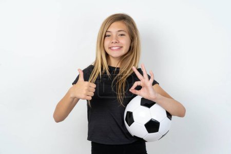 Téléchargez les photos : Adolescente portant des vêtements de sport tenant un ballon de football sur un mur blanc souriant et regardant heureux, insouciant et positif, geste de victoire ou de paix avec une main - en image libre de droit