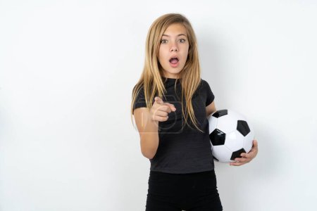 Photo for Shocked beautiful caucasian teen girl wearing sportswear holding a football ball over white wall points at you with stunned expression - Royalty Free Image