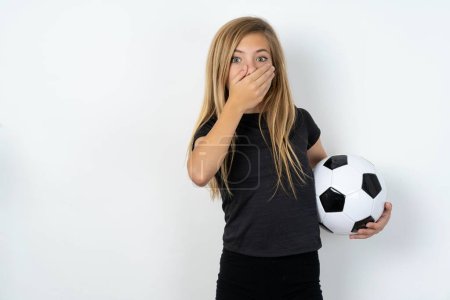 Foto de Aturdido hermosa chica adolescente caucásica usando ropa deportiva sosteniendo una pelota de fútbol sobre la pared blanca cubre ambas manos en la boca, temores de algo asombroso - Imagen libre de derechos