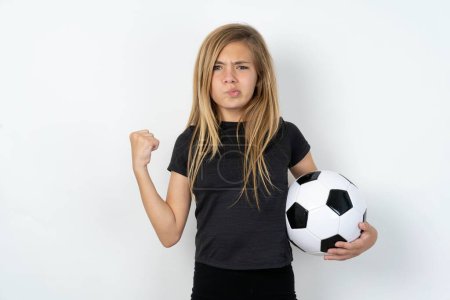 Photo for Irritated teen girl wearing sportswear holding a football ball over white wall blows cheeks with anger and raises clenched fists expresses rage and aggressive emotions. Furious model - Royalty Free Image
