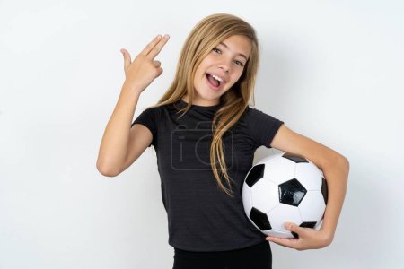 Photo for Teen girl wearing sportswear holding a football ball over white wall foolishness around shoots in temple with fingers makes suicide gesture. Funny model makes finger gun pistol - Royalty Free Image