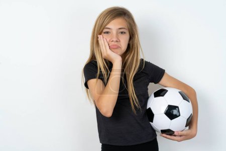 Photo for Sad lonely teen girl wearing sportswear holding a football ball over white wall touches cheek with hand bites lower lip and gazes with displeasure. Bad emotions - Royalty Free Image