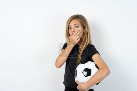 Téléchargez les photos : Adolescent fille portant des vêtements de sport tenant un ballon de football sur blanc couvre la bouche et regarde avec émerveillement à la caméra, ne peut pas croire rumeurs inattendues. - en image libre de droit