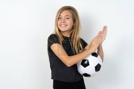 Téléchargez les photos : Surpris émotionnel adolescent fille portant des vêtements de sport tenant un ballon de football sur le mur blanc frotte les paumes et regarde la caméra avec incrédulité - en image libre de droit