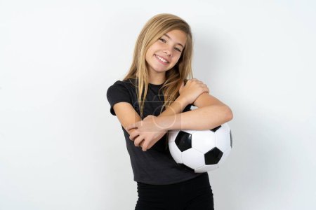 Téléchargez les photos : Charmante adolescente heureuse portant des vêtements de sport tenant un ballon de football sur un mur blanc embrasse son propre corps, se sent agréablement poses confortables. Concept de tendresse et d'estime de soi - en image libre de droit