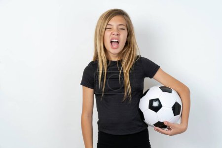 Foto de Adolescente con ropa deportiva sosteniendo una pelota de fútbol sobre bostezos pared blanca con la boca abierta se levanta. Rutina diaria - Imagen libre de derechos