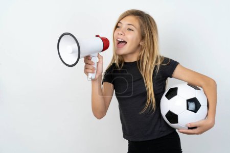 Lustiges Teenie-Mädchen in Sportkleidung hält einen Fußballball über die weiße Wand und schreit in Megafon. Mock up copy space.teen Mädchen in Sportbekleidung hält einen Fußball über weiße Wand 
