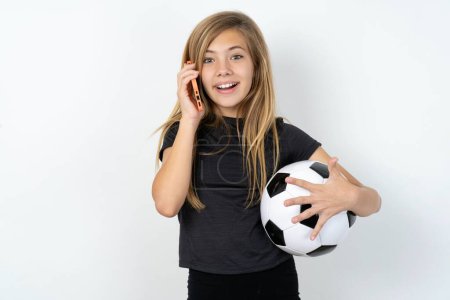Photo for Smiling teen girl wearing sportswear holding a football ball over white wall talks via cellphone, enjoys pleasant great conversation. People, technology, communication concept - Royalty Free Image
