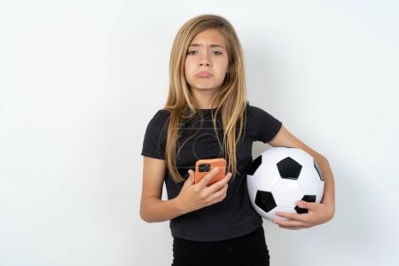 Foto de Muchacha adolescente insatisfecha con ropa deportiva sosteniendo una pelota de fútbol sobre la pared blanca utiliza la aplicación de software móvil y navega información en Internet, sostiene la mano móvil moderna - Imagen libre de derechos