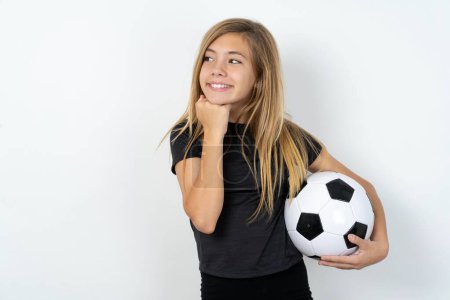 Foto de Adolescente chica usando ropa deportiva celebración de una pelota de fútbol sobre blanco pared sostiene las manos bajo la barbilla, contento de escuchar palabras conmovedoras de extraño - Imagen libre de derechos