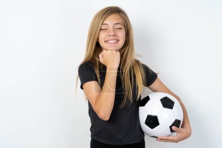 Foto de Chica adolescente alegre usando ropa deportiva sosteniendo una pelota de fútbol sobre la pared blanca tiene tímida expresión satisfecha, sonríe ampliamente, muestra dientes blancos, las emociones de la gente - Imagen libre de derechos