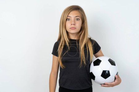 Foto de Jovencita aturdida usando ropa deportiva sosteniendo una pelota de fútbol sobre la pared blanca mira las reacciones en noticias impactantes. Chica asombrada aguanta la respiración - Imagen libre de derechos